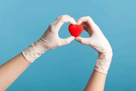Profile side view closeup of human hand in white surgical gloves holding smal red heart shape in hand. indoor, studio shot, isolated on blue background.の写真素材