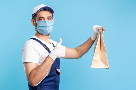 Portrait of satisfied handyman in overalls, mask and gloves holding ordered parcel, showing thumbs...