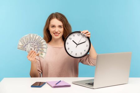 Time to make money profit! Positive happy woman sitting at workplace, holding clock and dollar banknotes, looking at camera with charming toothy smile. indoor studio shot isolated on blue backgroundの写真素材