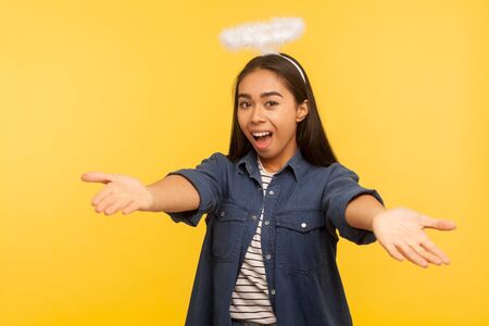 Come into my arms! Portrait of angelic kind generous girl with nimbus keeping hands wide open to embrace, giving warm welcome, greeting and sharing love. studio shot isolated on yellow backgroundの写真素材