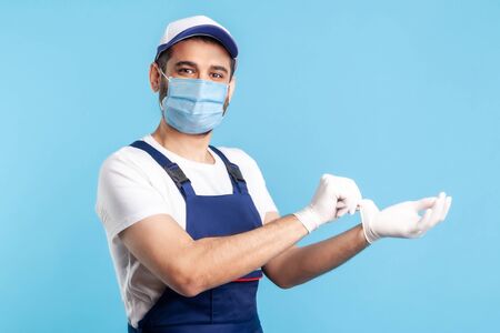 handyman in overalls and mask showing hand with surgical gloves, smiling to camera. Safety clothes, protection and hygiene in service industry, courier delivery, housekeeping maintenance. indoor.の写真素材