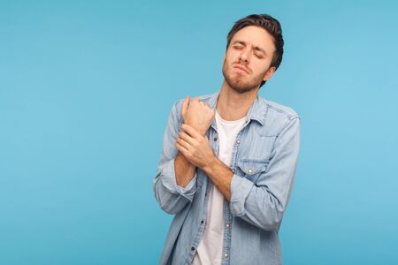 Portrait of office worker man in denim shirt touching painful hand, suffering trauma, sprain wrist, feeling ache of carpal tunnel syndrome. indoor studio shot isolated on blue backgroundの写真素材