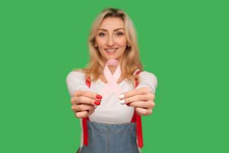 Beautiful optimistic adult woman in stylish denim overalls showing pink ribbon, symbol of oncological disease prevention. indoor studio shot isolated on green backgroundの写真素材