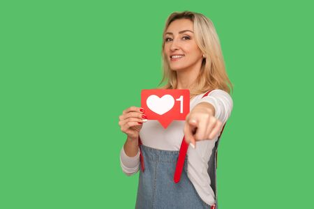 Portrait of lovely cheerful adult woman holding network heart icon and pointing to camera, showing emoji button. studio shot isolated on green backgroundの写真素材