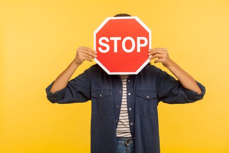 Attention, prohibited way! Unknown person in denim shirt hiding face behind Stop symbol, red traffic sign warning of restricted access, banned service. indoor studio shot isolated on yellow backgroundの写真素材