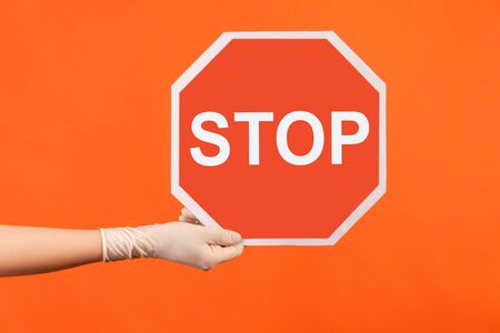 Profile side view closeup of human hand in white surgical gloves holding and showing Stop sign. indoor, studio shot, isolated on orange background.の写真素材
