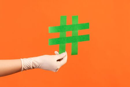 Profile side view closeup of human hand in white surgical gloves holding green hashtag. indoor, studio shot, isolated on orange background.の写真素材
