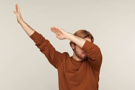 Dabbing movement. Portrait of optimistic carefree man with beard wearing sweatshirt standing in dab dance pose, celebrating success, popular internet meme. studio shot isolated on gray backgroundの写真素材