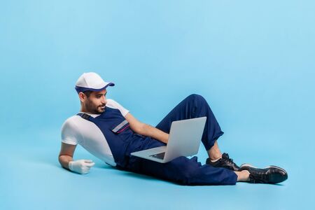 Full length handsome expert handyman in uniform lying sexy posing on camera, reading message on laptop. Profession of online customer support, contact center of repair service. studio shot isolatedの写真素材