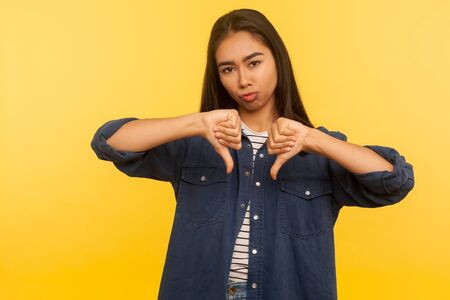 Awful result! Portrait of dissatisfied girl in denim shirt showing thumbs down, disapproval sign,...