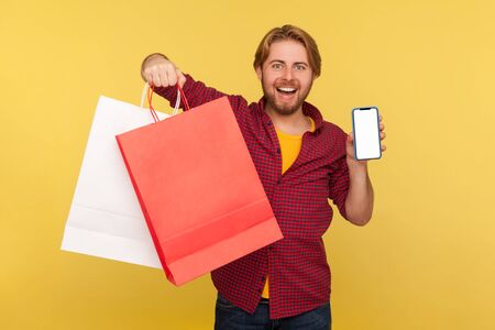 Happy handsome shopper guy in checkered shirt holding shopping bags and mobile phone blank mock up...