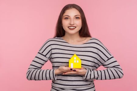 Real estate purchase. Portrait of happy young woman in striped sweatshirt holding paper house and looking at camera with toothy smile, dreaming of home. indoor studio shot isolated on pink backgroundの写真素材