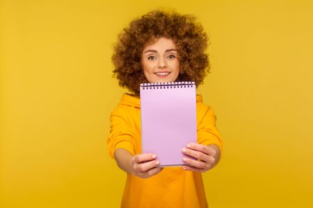 Portrait of joyful happy student, curly-haired woman in urban style hoodie showing blank notebook, advertising area on paper, copy space for to-do list, idea. studio shot isolated on yellow backgroundの写真素材