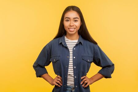 Portrait of cheerful happy girl in denim shirt looking at camera with toothy smile and holding hands on hips, confident expression, optimistic view. indoor studio shot isolated on yellow backgroundの写真素材