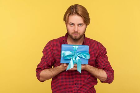 Handsome stylish bearded guy in checkered shirt holding elegant gift box, looking right into camera, greeting happy birthday, congratulating on anniversary. studio shot isolated on yellow backgroundの写真素材