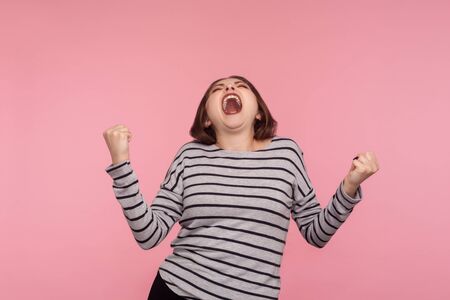 Triumphal victory! Portrait of extremely happy, overjoyed woman in striped sweatshirt yelling for joy with raised fists, celebrating success, euphoria from win. studio shot isolated on pink backgroundの写真素材