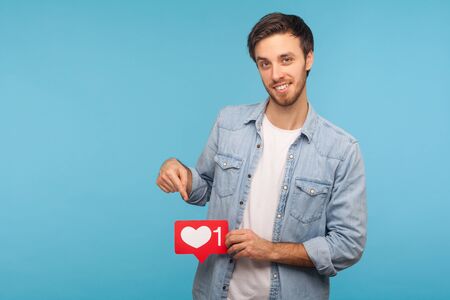 Click Like and subscribe! Portrait of handsome blogger man in denim shirt pointing at social media heart button, emoji counter, follower notification. indoor studio shot isolated on blue backgroundの写真素材