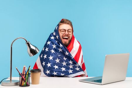 Extremely happy funny man employee sitting wrapped in American flag and shouting for joy in office workplace, celebrating labor day or US Independence day 4th of july, government employment support.の写真素材