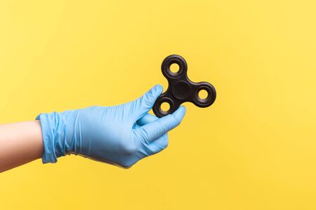 Profile side view closeup of human hand in blue surgical gloves holding black fidget spinner. indoor, studio shot, isolated on yellow background.の写真素材