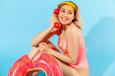 Portrait of charming happy attractive woman in swimsuit sitting with rubber ring and talking on...