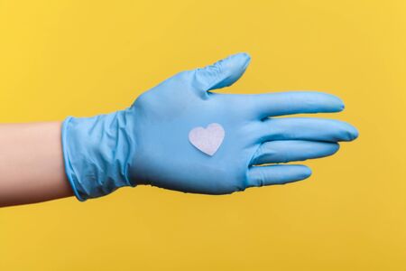 Profile side view closeup of human hand in blue surgical gloves holding small pink heart shape in...
