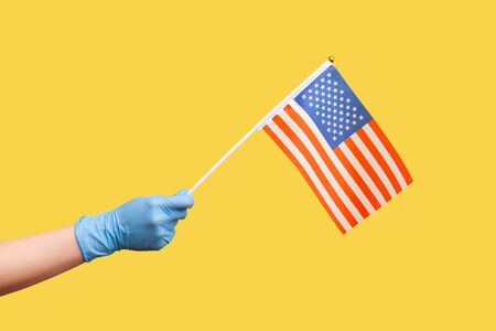 Profile side view closeup of human hand in blue surgical gloves holding USA national flag. United states of America. National independance day - 4th july. indoor, isolated on yellow background.の写真素材