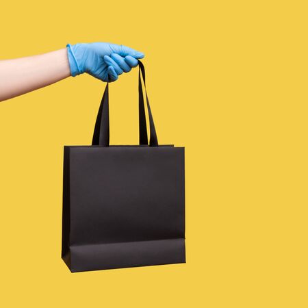 Profile side view closeup of human hand in blue surgical gloves holding and showing black shopping bag. indoor, studio shot, isolated on yellow background.の写真素材