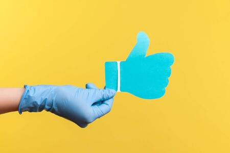 Profile side view closeup of human hand in blue surgical gloves holding and showing like or thumbs up paper shape sign. indoor, studio shot, isolated on yellow background.の写真素材
