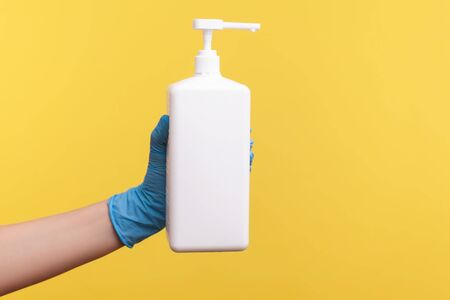 Profile side view closeup of human hand in blue surgical gloves holding and showing sanitizer, antiseptic or anti bacterial soap . indoor, studio shot, isolated on yellow background.の写真素材