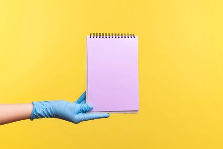 Profile side view closeup of human hand in blue surgical gloves holding purple notepad in hand and holding empty paper. indoor, studio shot, isolated on yellow background.の写真素材