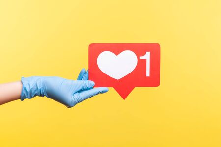 Profile side view closeup of human hand in blue surgical gloves holding social media like stick. indoor, studio shot, isolated on yellow background.の写真素材