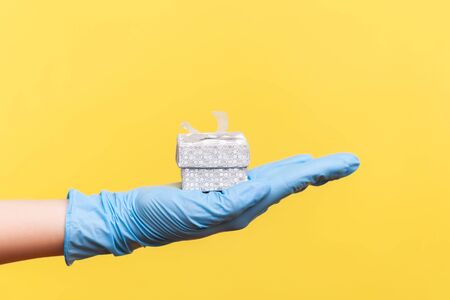 Profile side view closeup of human hand in blue surgical gloves holding small gift box. sharing, giving wedding proposal, or delivery concept. indoor, studio shot, isolated on yellow background.の写真素材
