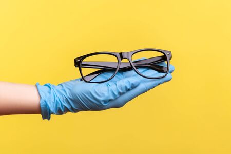 Profile side view closeup of human hand in blue surgical gloves holding and giving black eyeglasses frame. indoor, studio shot, isolated on yellow background.の写真素材