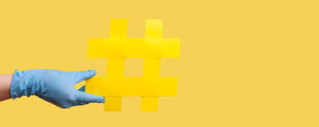 Profile side view closeup of human hand in blue surgical gloves holding yellow hashtag. indoor, studio shot, isolated on yellow background.の写真素材