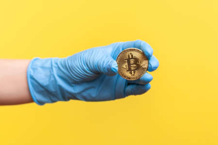 Profile side view closeup of human hand in blue surgical gloves holding and showing bitcoin symbol in hand. indoor, studio shot, isolated on yellow background.の写真素材