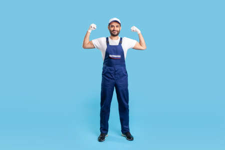 Full length cheerful handyman in overalls, cap and protective gloves standing, showing biceps, demonstrating strength. Profession of service industry, loader or building worker. studio shot isolatedの写真素材