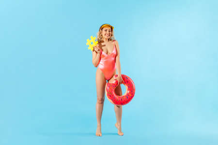 Portrait of lovely carefree beautiful woman in swimsuit holding rubber ring and sweet lollipop candy, enjoying rest on tropical beach, vacation resort. indoor studio shot isolated on blue backgroundの写真素材