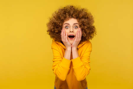 Take sweet dessert! Portrait of happy curly-haired young woman in urban style hoodie giving delicious sugary donut and smiling genuinely to camera. indoor studio shot isolated on yellow backgroundの写真素材