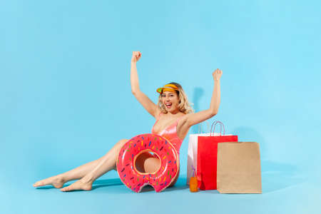 Portrait of enthusiastic overjoyed girl in swimsuit sitting with rubber ring and shopping bags,...