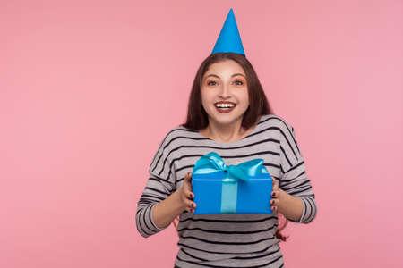 Happy birthday to me! Joyful optimistic woman with party cone hat smiling at camera and holding wrapped gift box, rejoicing present, holiday celebration. studio shot isolated on pink backgroundの写真素材