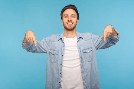Portrait of handsome happy man in stylish denim shirt smiling and pointing down, showing place for idea presentation, commercial text. indoor studio shot isolated on blue backgroundの写真素材