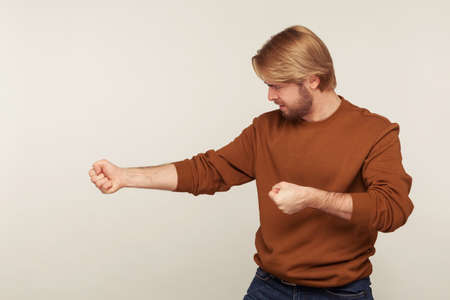 Portrait of strong purposeful man with beard in sweatshirt keeping fists up, holding invisible virtual rope and pulling object, striving hard with great effort. studio shot isolated on gray backgroundの写真素材