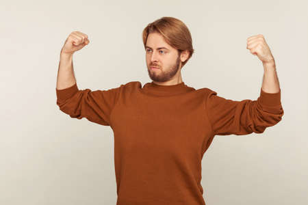 Portrait of confident proud bearded man in sweatshirt demonstrating power in his hands, showing...