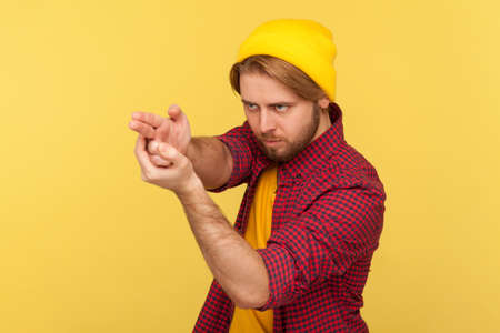 Dangerous hooligan hipster guy in beanie hat and checkered shirt pointing finger gun, threatening to kill, shooting with weapon gesture right in target. studio shot isolated on yellow backgroundの写真素材