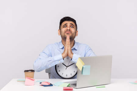 Unhappy man employee sitting in office workplace, holding clock looking imploring with prayer...