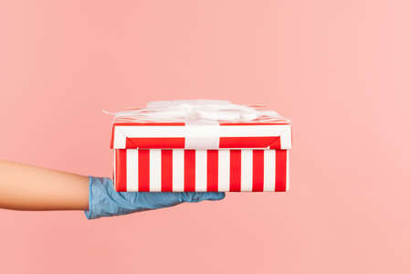 Profile side view closeup of human hand in blue surgical gloves holding striped red white gift box. indoor, studio shot, isolated on pink background.の写真素材