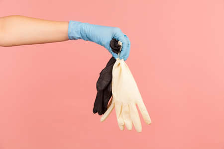 Profile side view closeup of human hand in blue surgical gloves holding white and black gloves. indoor, studio shot, isolated on pink background.の写真素材