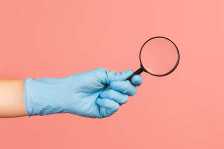 Profile side view closeup of human hand in blue surgical gloves holding magnify glass. indoor, studio shot, isolated on pink background.の素材 [FY310152729376]