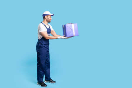 Take present! Full length professional courier in overalls and protective gloves giving wrapped box,...