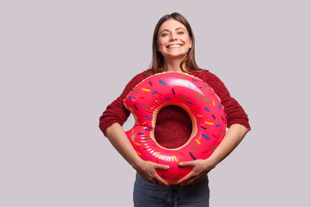 Optimistic cheerful girl holding rubber ring pink donut and looking at camera with toothy smile, enjoying summer vacation, rest at seaside, travel concept. studio shot isolated on gray backgroundの写真素材